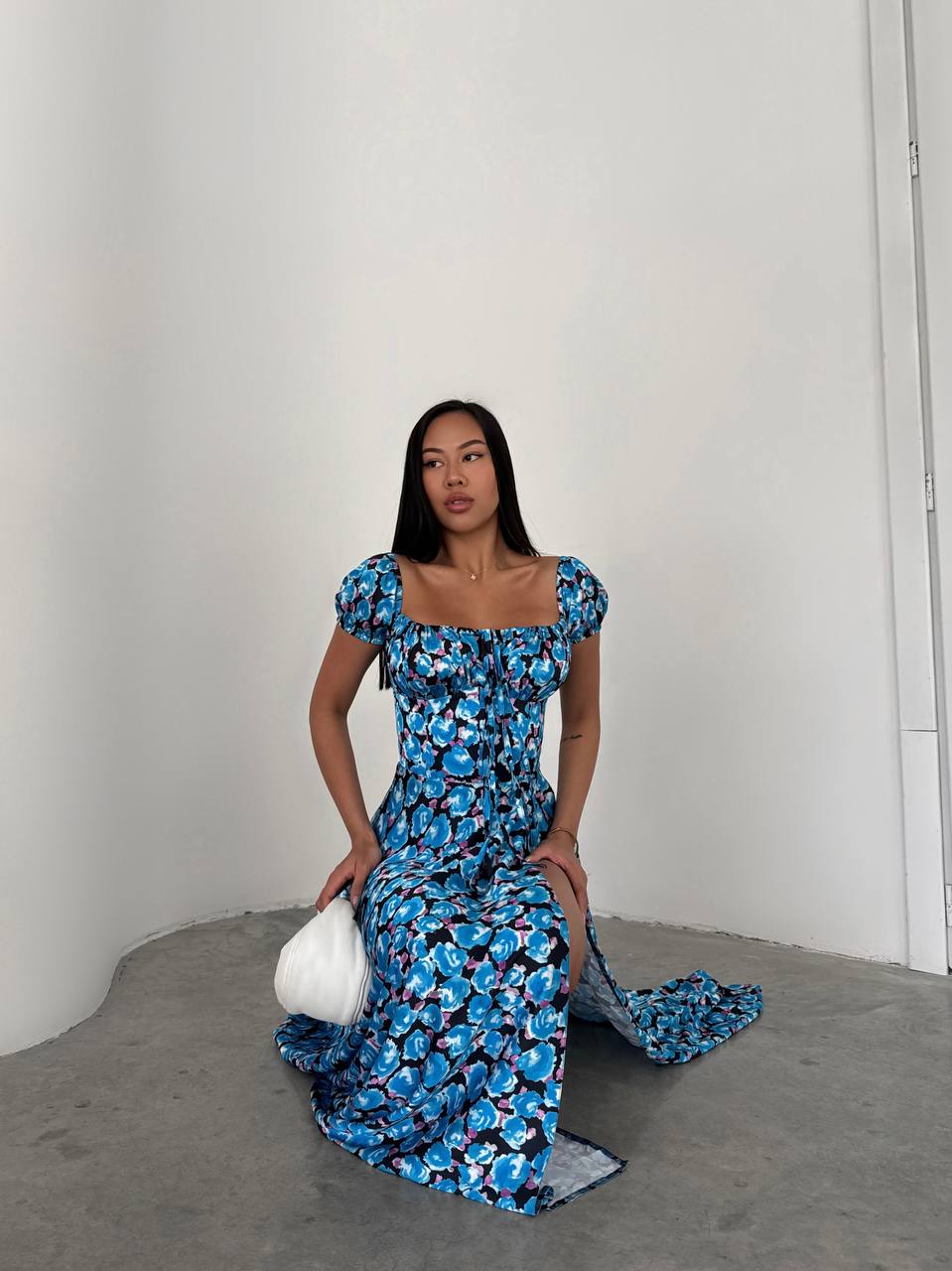 Woman in a blue floral dress holding a white clutch against a plain background