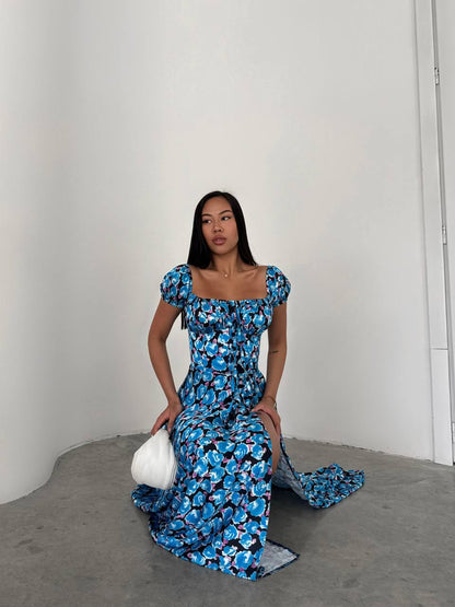Woman in a blue floral dress holding a white clutch against a plain background