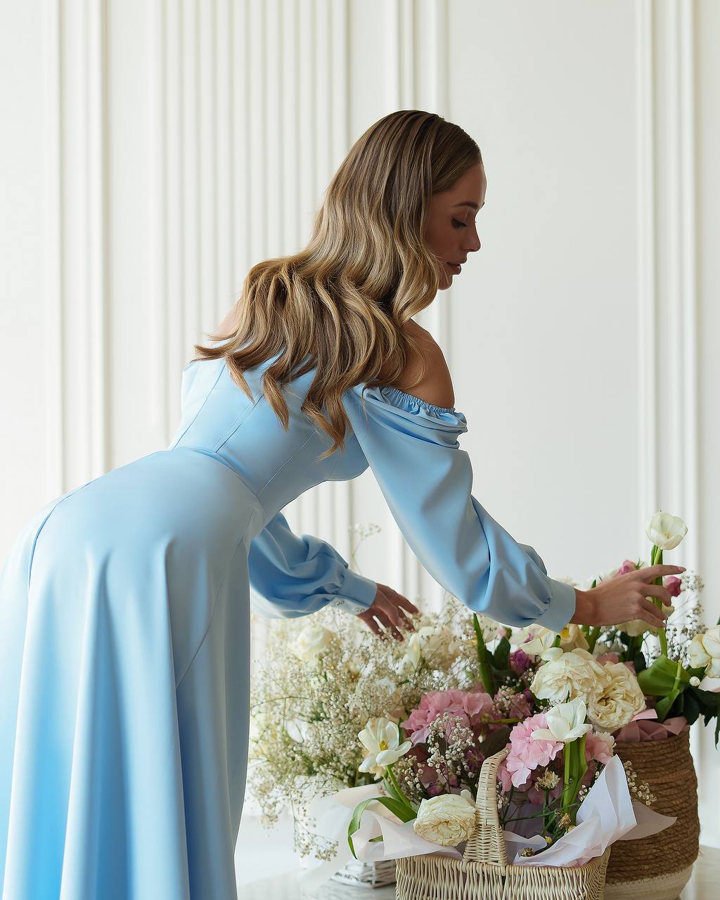 Woman in a light blue dress arranging flowers in a room with white walls.