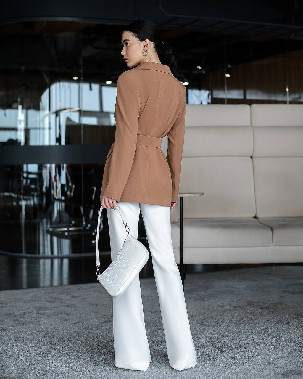Woman in a brown blazer and white pants holding a white bag in an indoor setting.