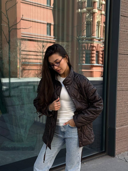 Woman wearing a brown puffer jacket, white shirt, and blue jeans standing outside a building.