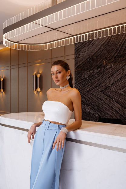 Woman in a white top and light blue pants leaning against a modern interior wall.