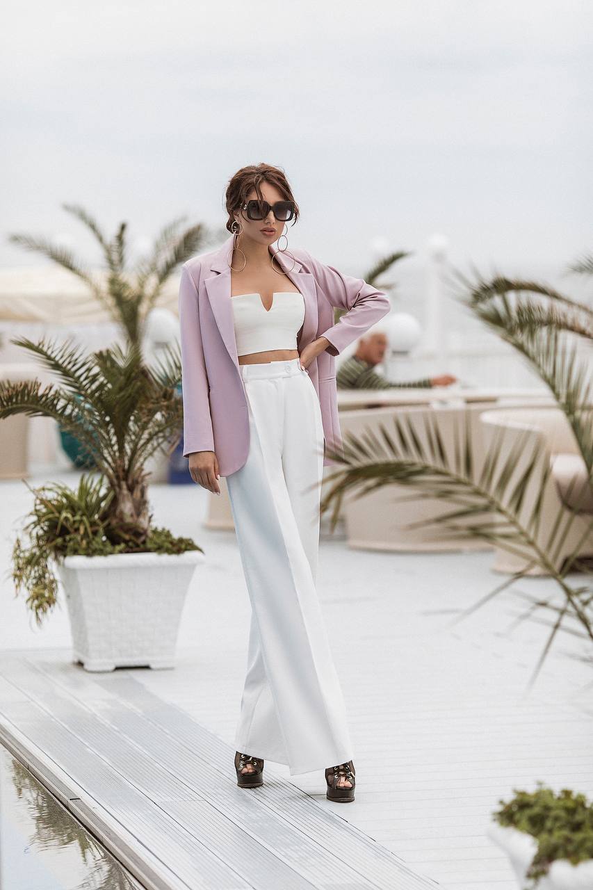 A woman stands on a white deck wearing a lavender blazer, white crop top, and white wide-leg trousers with black platform sandals. Palm plants and a blurred outdoor setting are visible in the background.