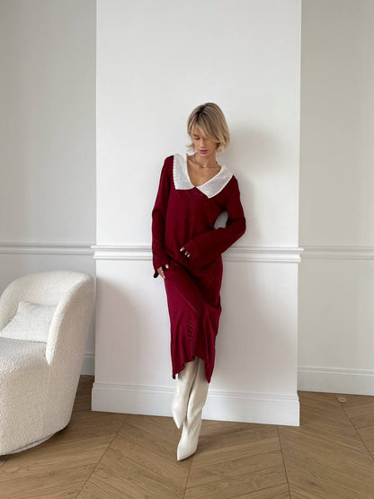 Woman in a burgundy dress with white collar standing against a white wall.