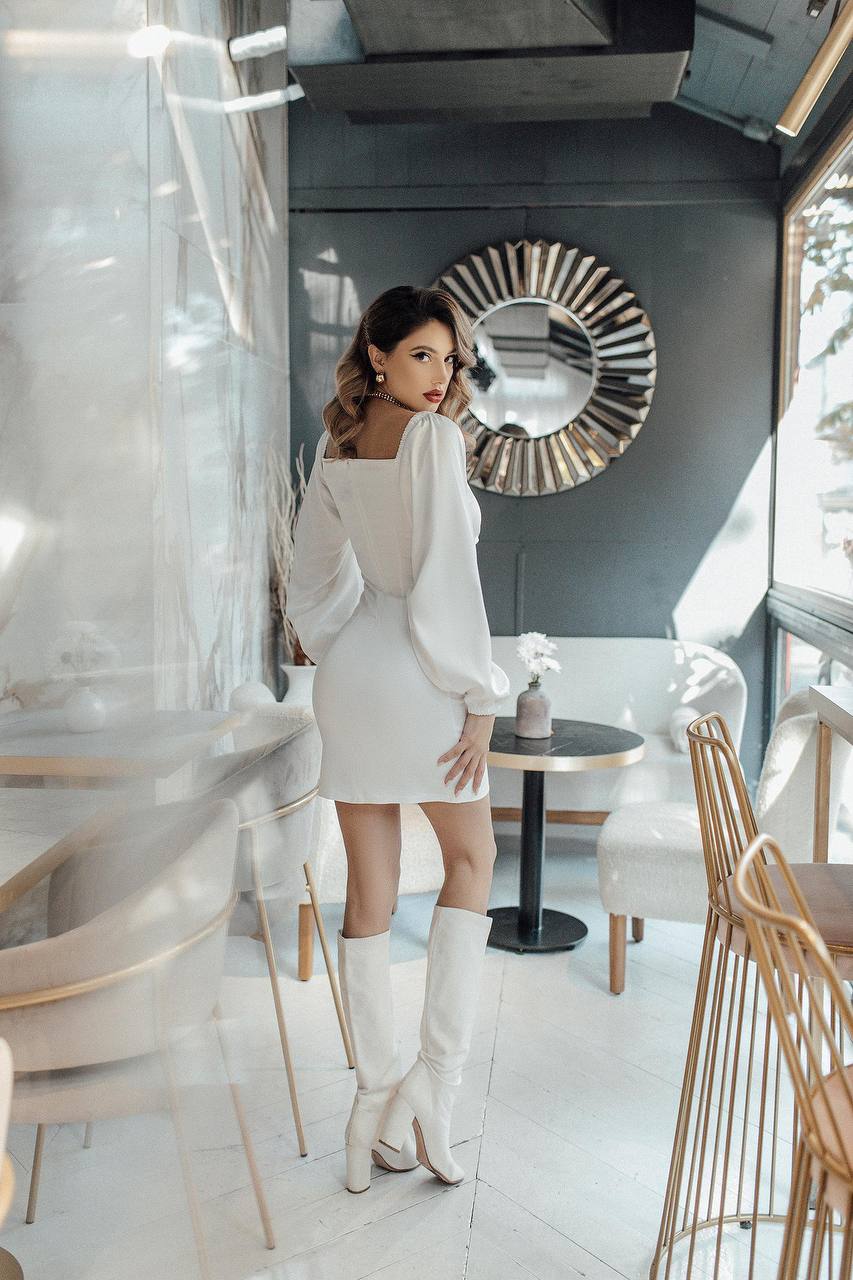 Woman in a white dress and knee-high boots standing in a modern interior setting.