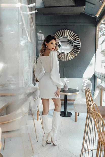 Woman in a white dress and knee-high boots standing in a modern interior setting.