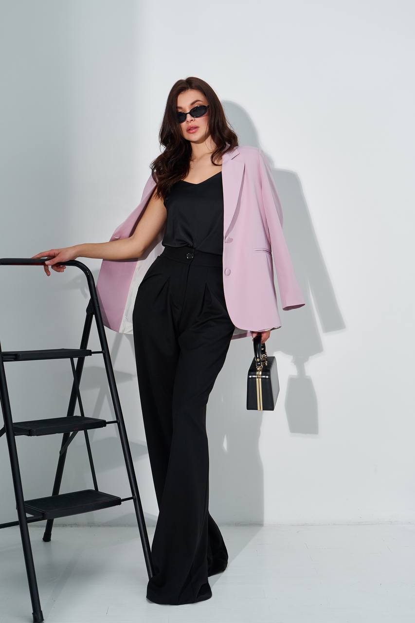A woman models a light pink blazer over a black top and black wide-leg trousers, holding a small black handbag with gold trim. She is standing next to a black metal step ladder against a white wall.
