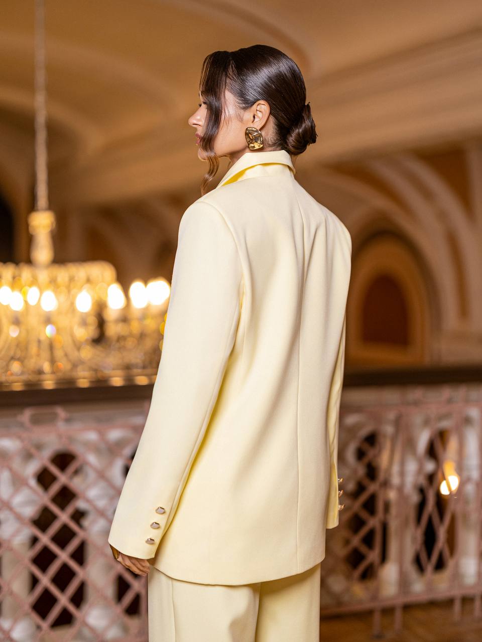 Woman in a light yellow suit standing in an ornate room with chandeliers.