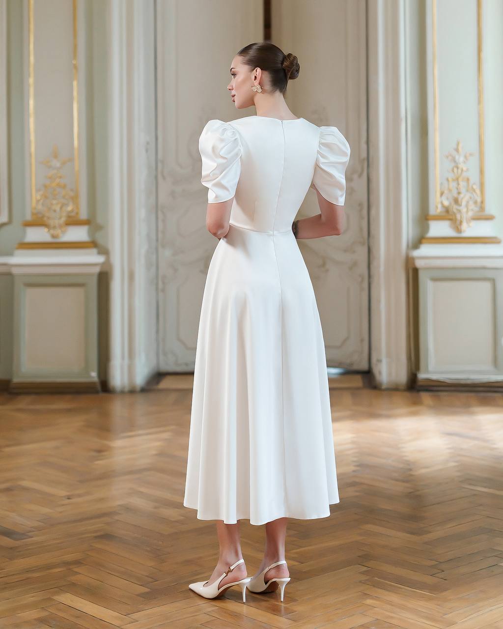 Woman in a white dress standing in an elegant room with wooden flooring and decorative walls.