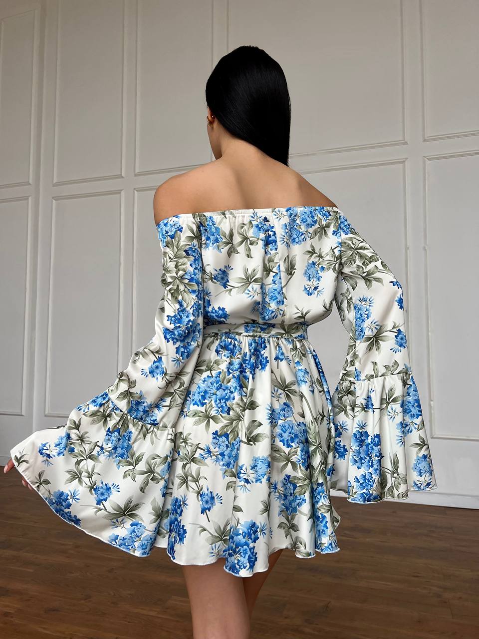 Woman wearing a floral off-shoulder dress with blue flowers against a neutral wall.
