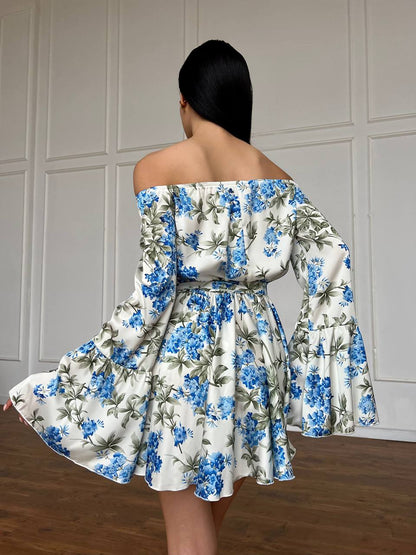 Woman wearing a floral off-shoulder dress with blue flowers against a neutral wall.