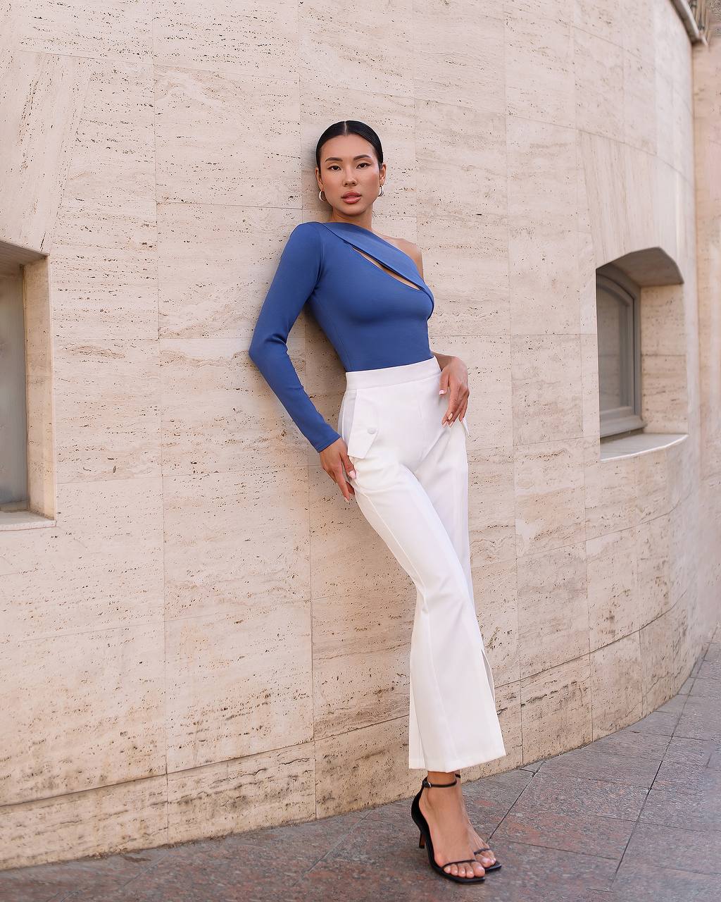 Woman in a blue top and white pants standing against a beige wall.