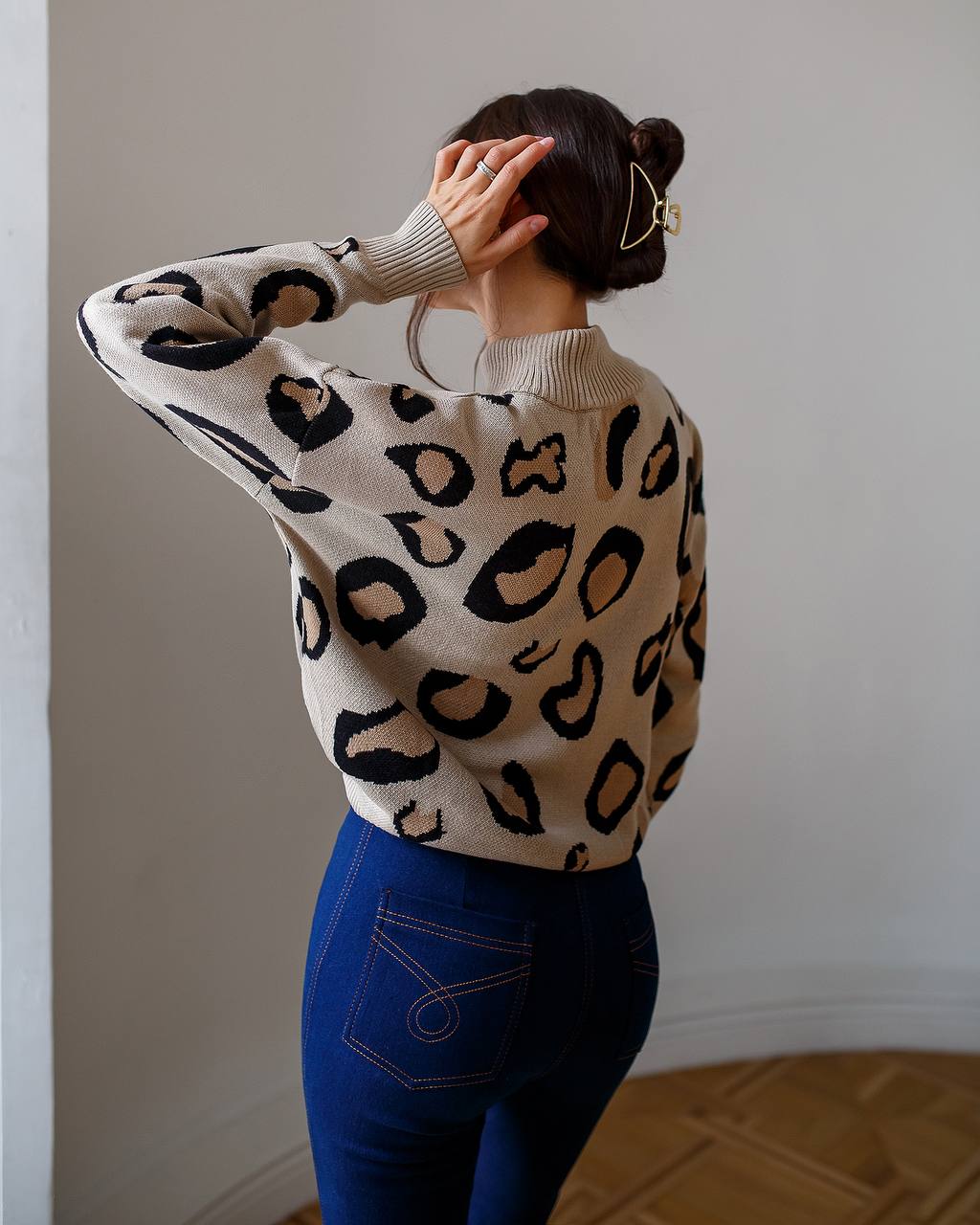 Person wearing a leopard print sweater and blue pants against a plain wall.