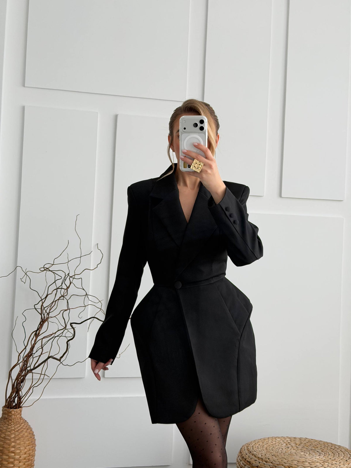 A woman takes a mirror selfie wearing a black tailored blazer-style dress with structured shoulders, a wrap-style front, and a belted waist. She is standing against a white paneled wall, with a woven basket vase and dried branches on the left and a round woven stool at the bottom right.