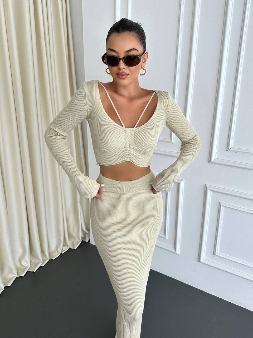 Woman in a cream-colored outfit posing against a white paneled wall.