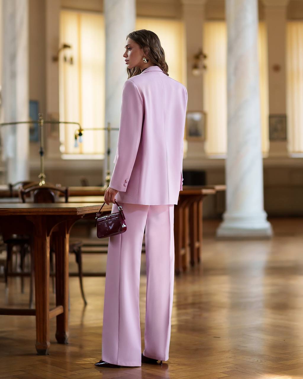 Woman in a pink suit standing in a room with wooden tables and columns.