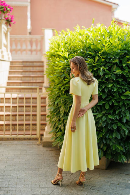 A woman wearing a pale yellow short-sleeve midi dress and brown strappy heels, standing outdoors in front of a large green hedge and stone stairs.