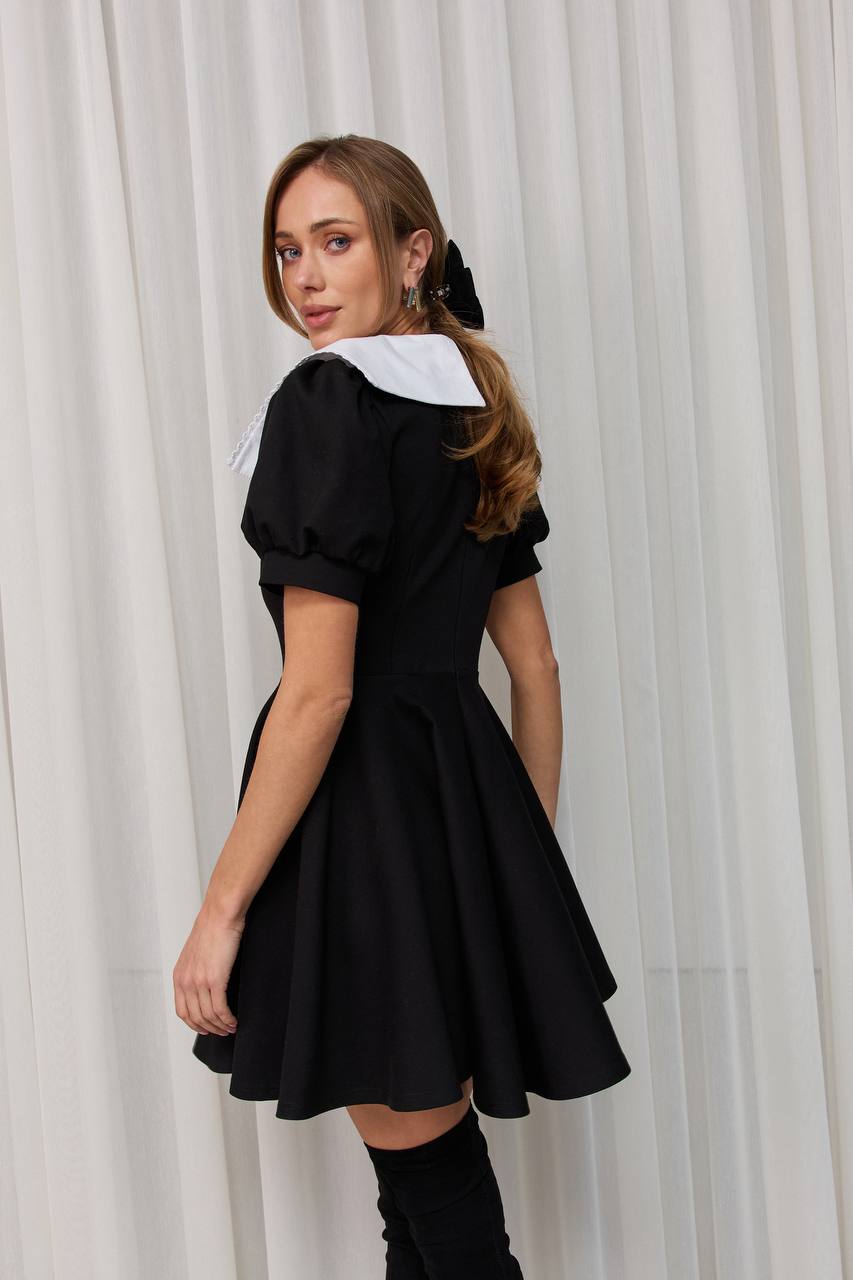 Woman wearing a black dress with a white collar against a white curtain background