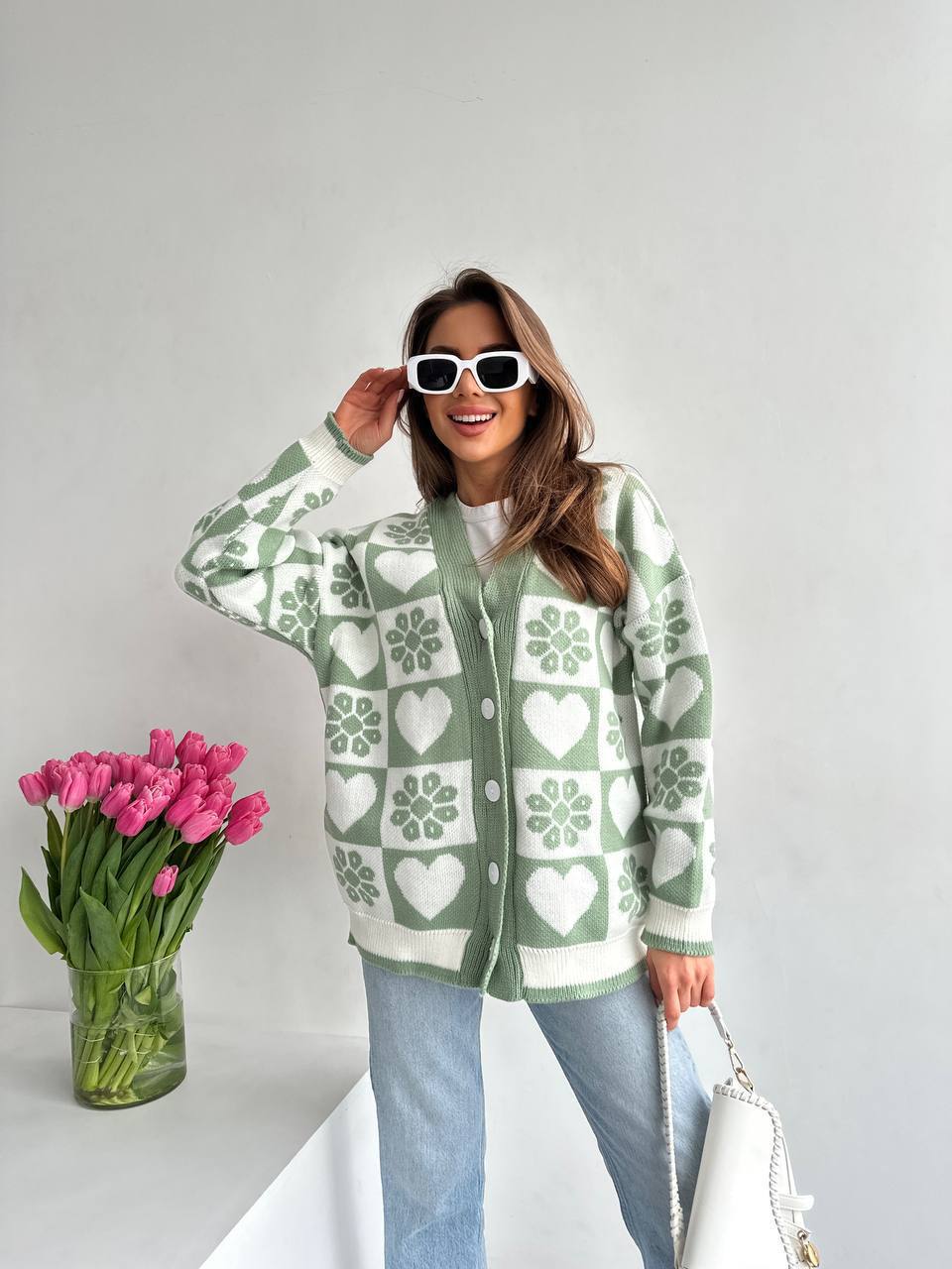 Woman wearing a green and white patterned cardigan with pink tulips and sunglasses on a light gray background
