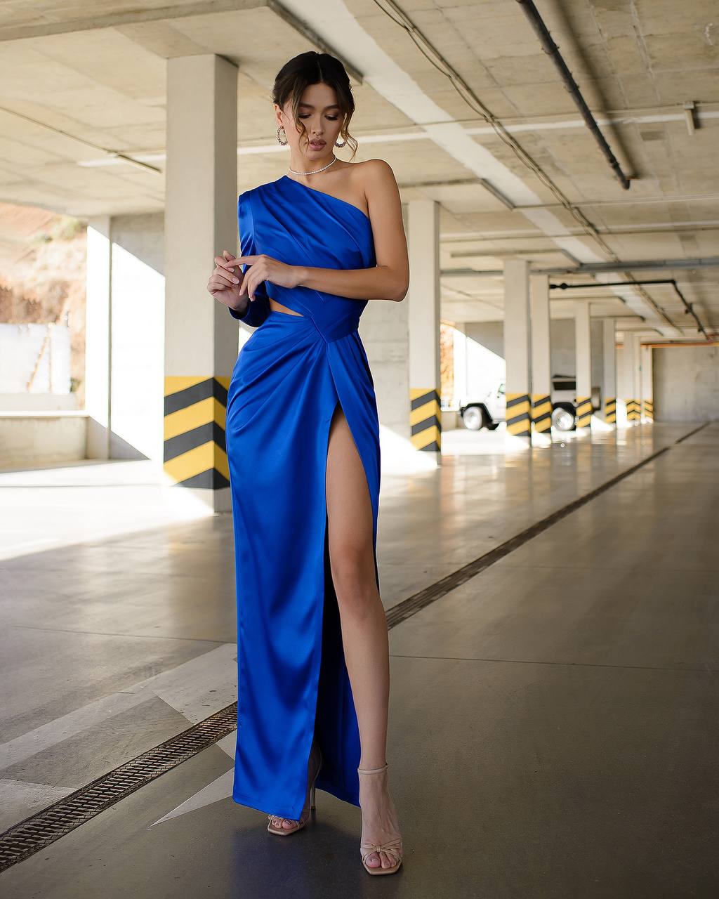 Woman in a blue one-shoulder dress with a high slit standing in an underground parking lot.