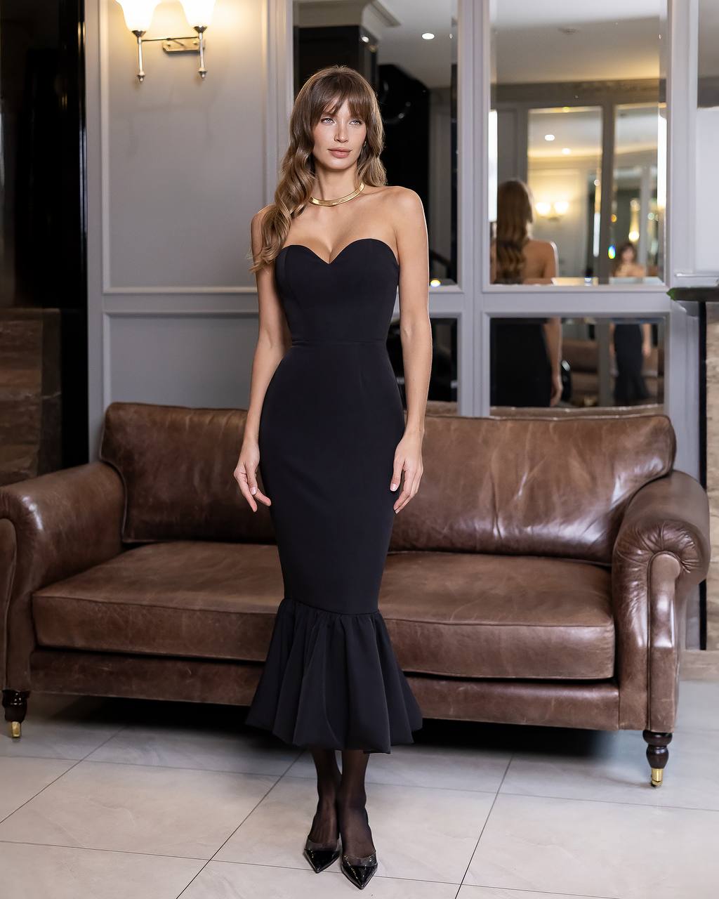 Woman in a black dress standing in a room with a brown leather sofa.