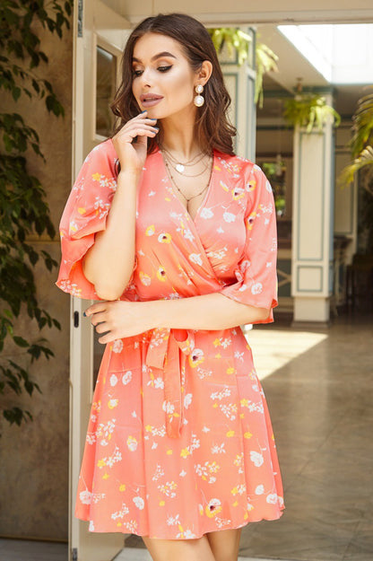 Woman wearing a coral floral dress standing in a sunlit room with plants.