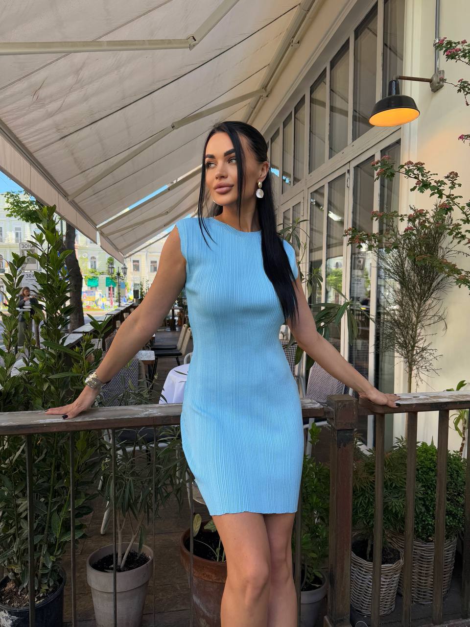 Woman in a light blue dress standing on a balcony with plants and outdoor furniture.