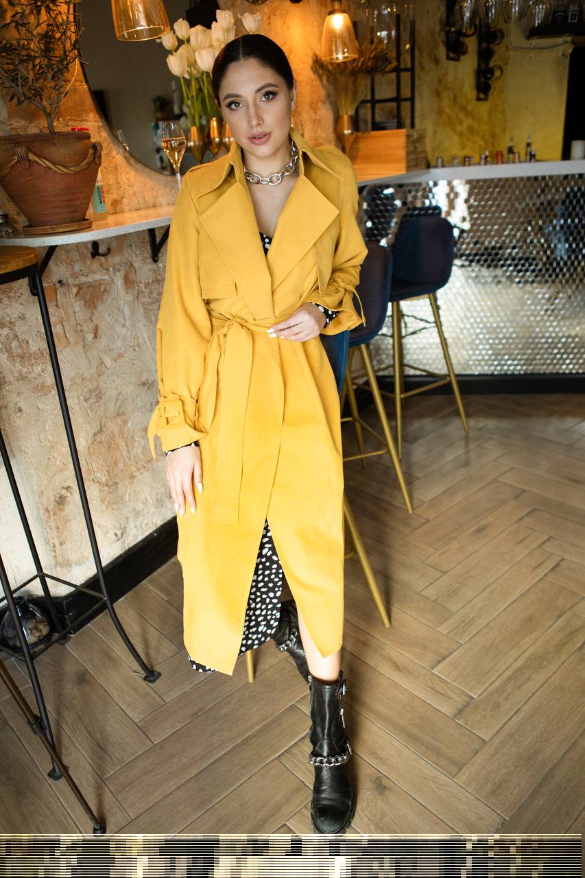Woman in a yellow trench coat standing in a stylish interior setting.