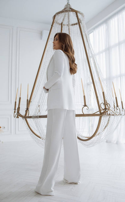 Woman in a white outfit standing in a bright room with large windows and decorative elements.