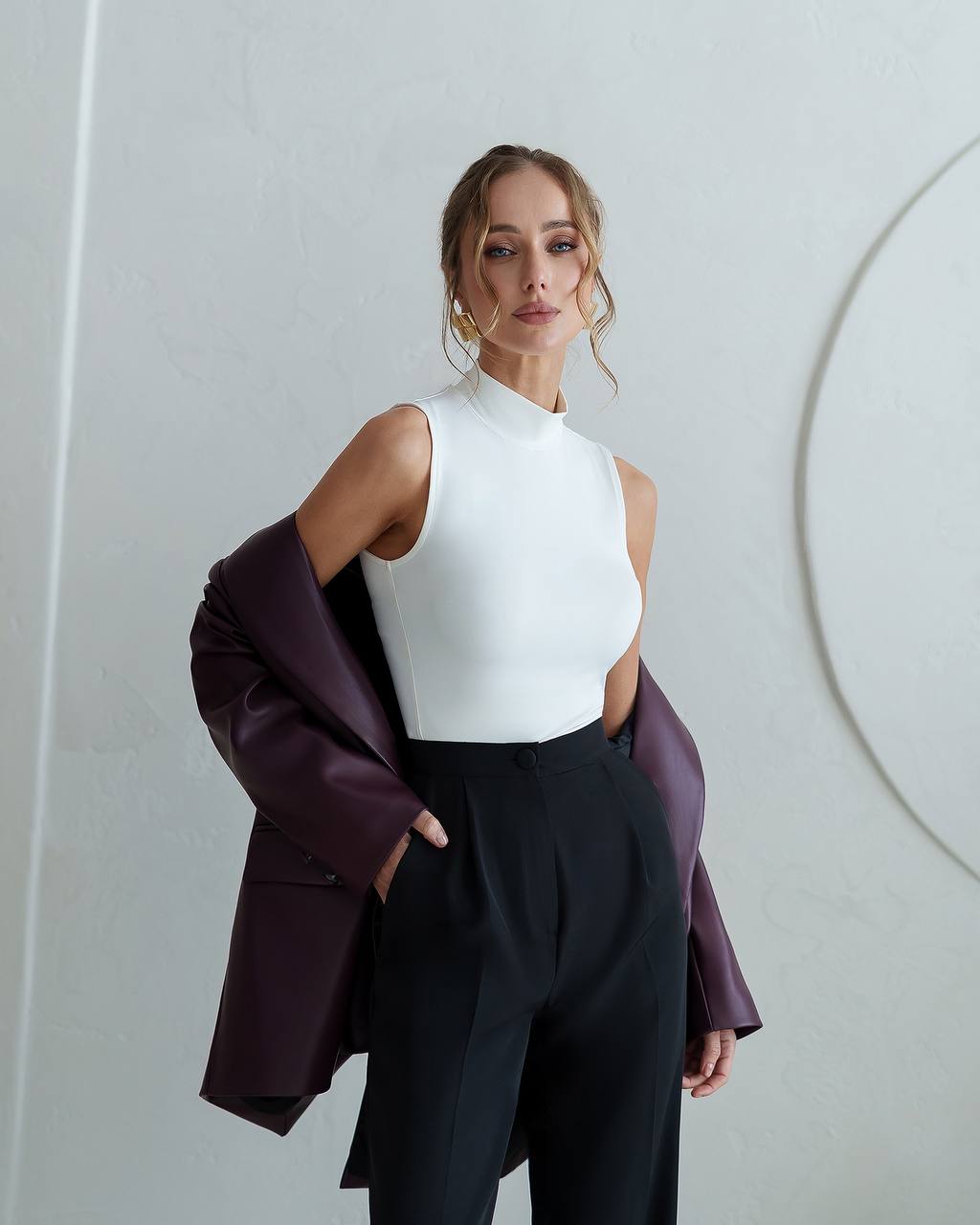Woman wearing a white sleeveless top and black pants with a purple coat against a light gray background