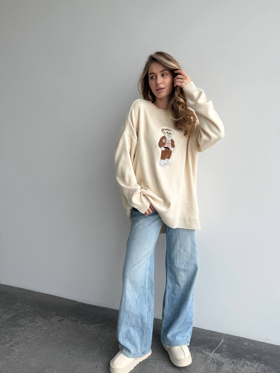 Woman wearing a cream sweatshirt with a graphic design and light blue jeans against a gray wall.