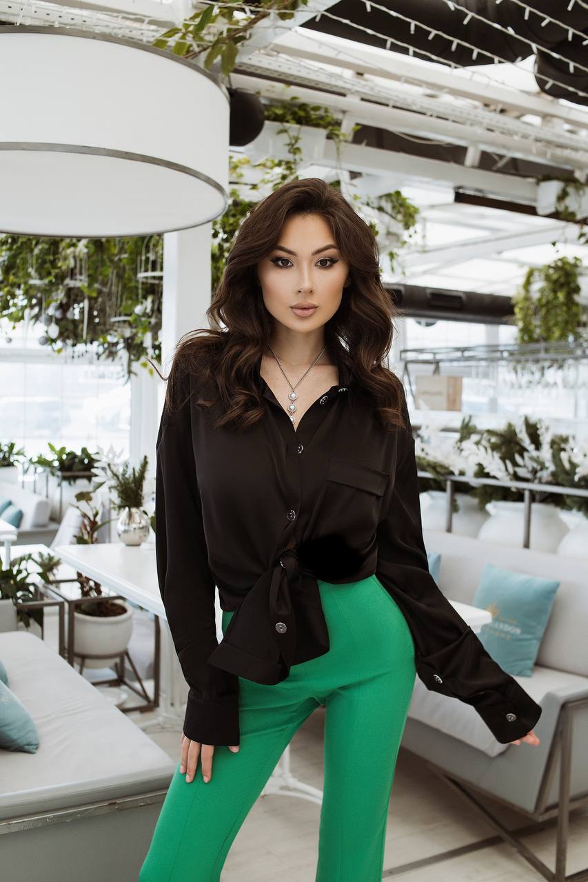 Woman in black top and green pants standing in an outdoor setting with white furniture and plants.
