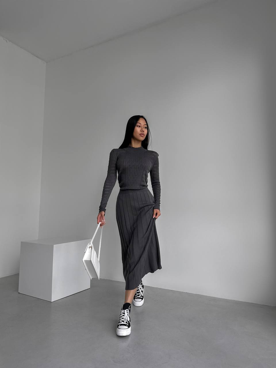 Woman in a gray outfit with black sneakers holding a white bag in a minimalistic room.