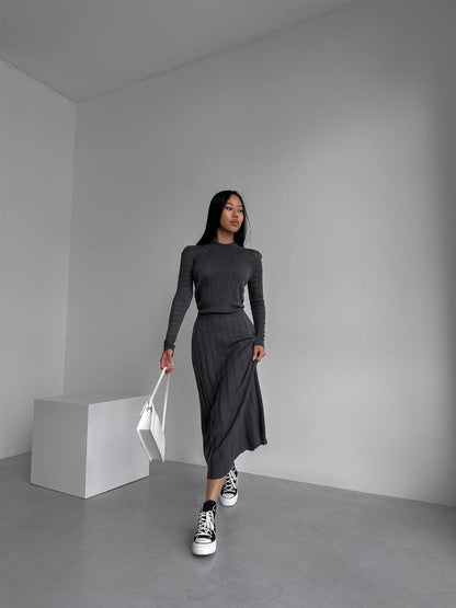 Woman in a gray outfit with black sneakers holding a white bag in a minimalistic room.