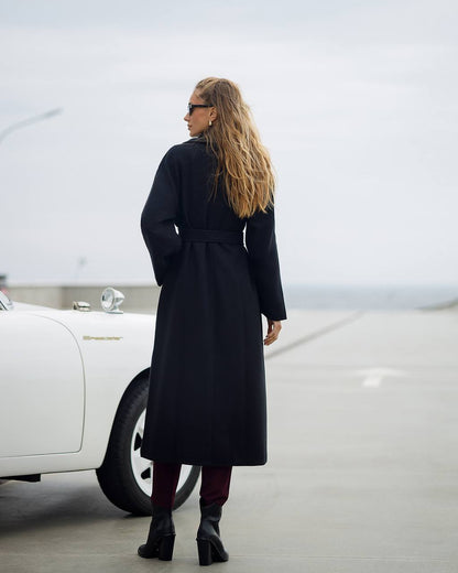 Woman in a long black coat standing next to a white car on a road.