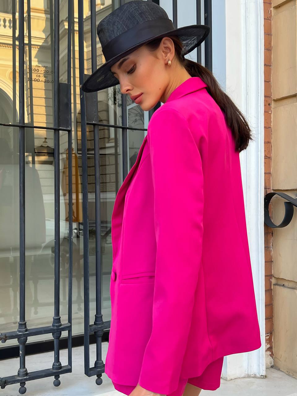 Person wearing a bright pink blazer and black hat in an urban setting.
