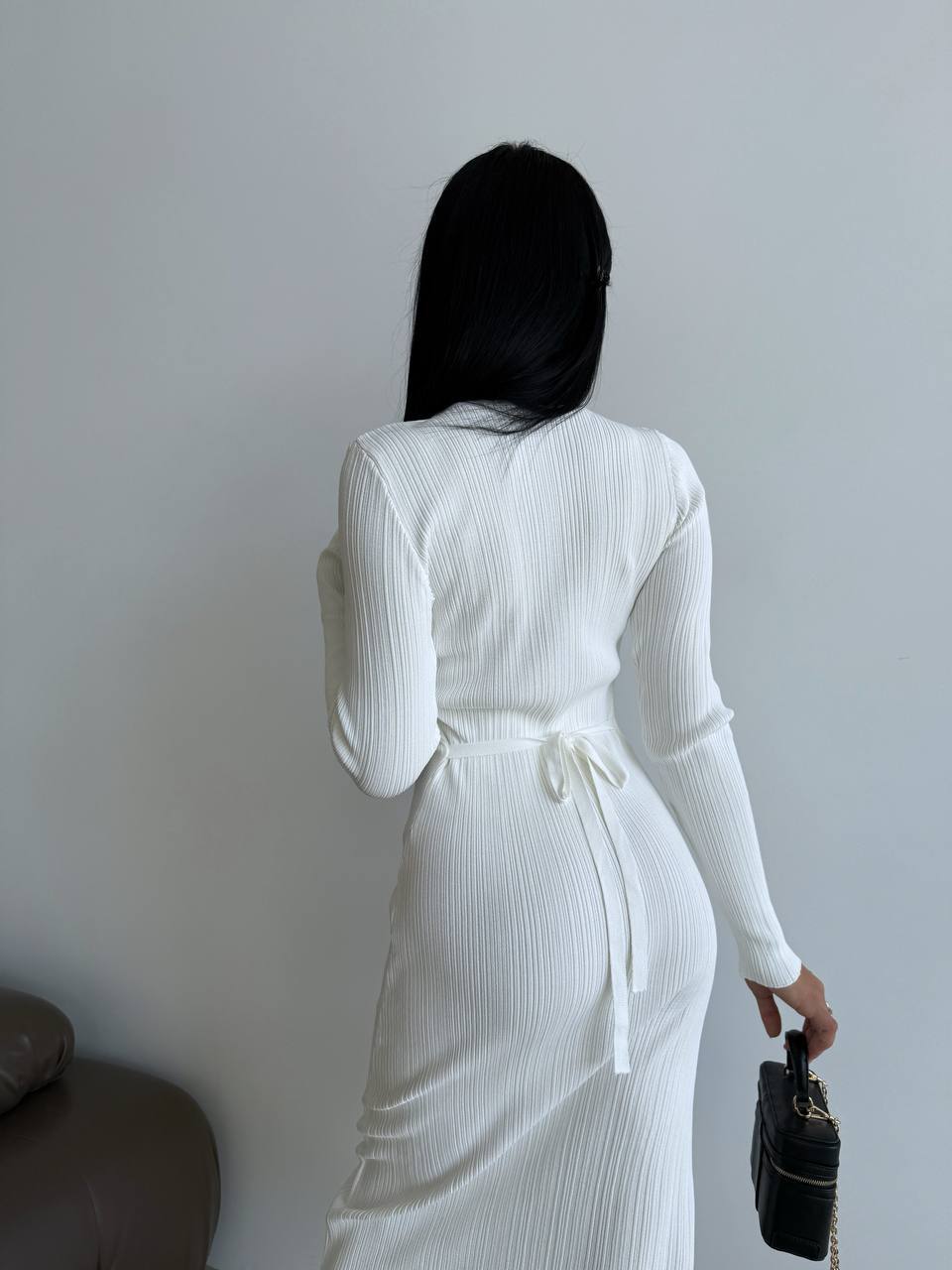 Person wearing a white ribbed dress with a belt, standing against a plain wall.
