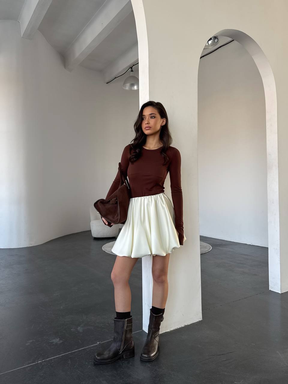 Woman in a brown top and white skirt standing in a minimalistic room.