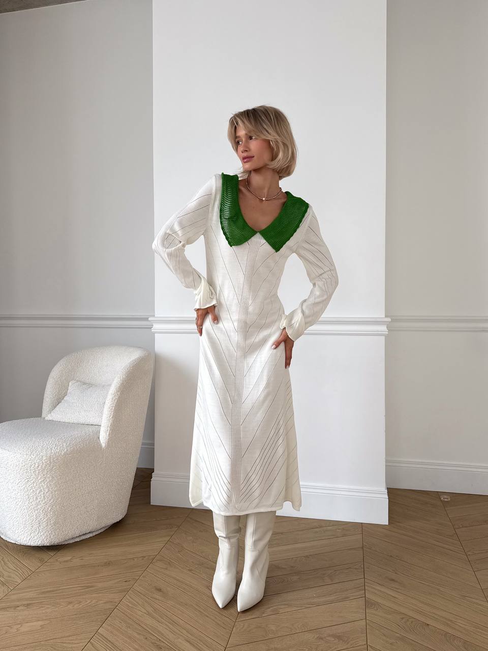 Woman in a white dress with green neckline standing in a room with a white chair and light-colored floor.