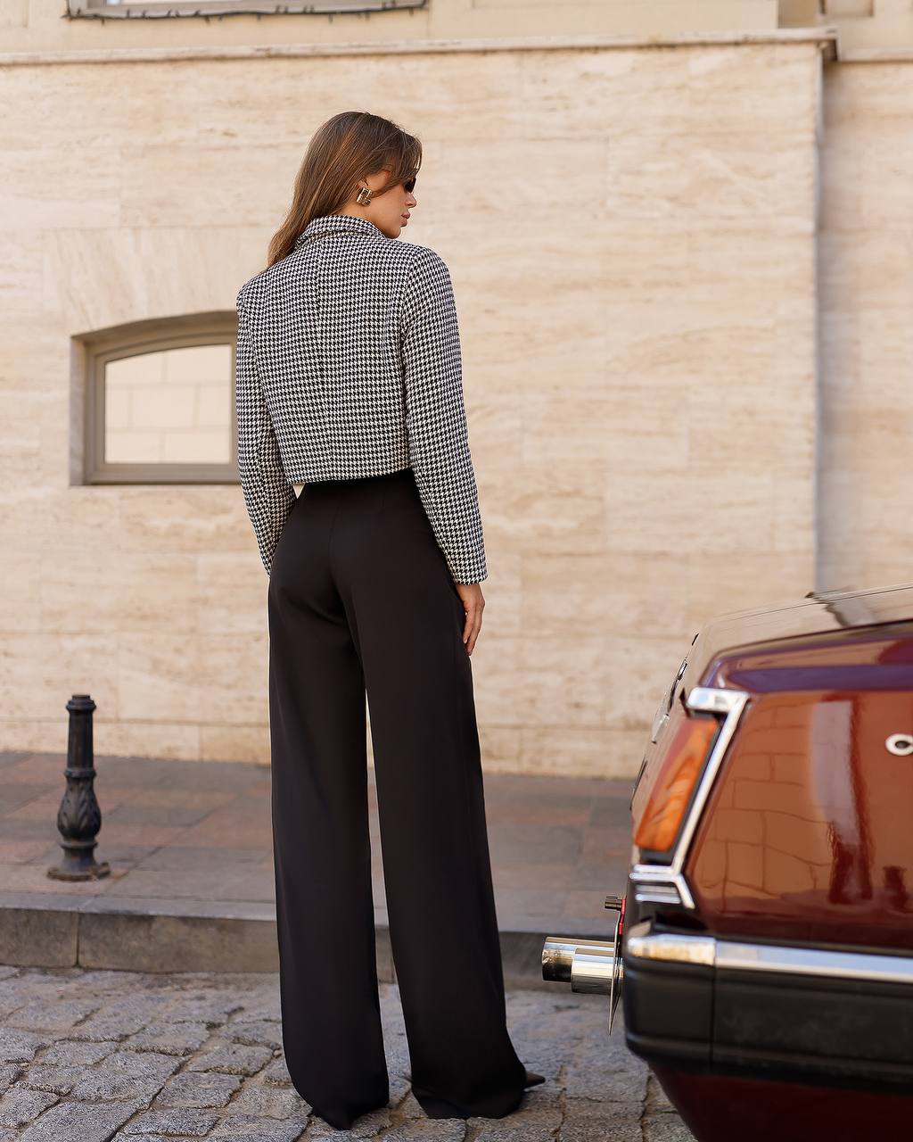 Woman in a checkered shirt and black pants standing next to a vintage car on a street.