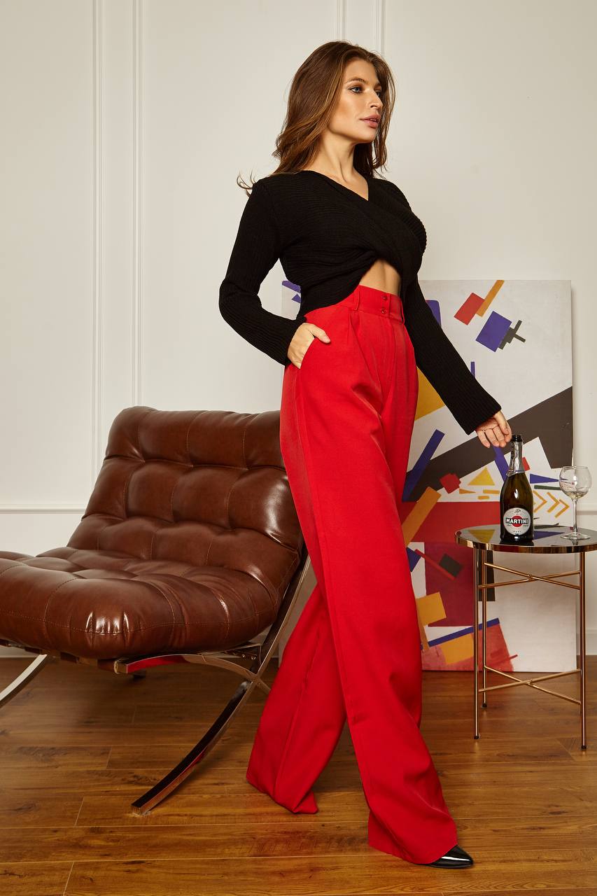 Woman in black top and red pants standing in a room with a brown leather chair and colorful abstract painting.