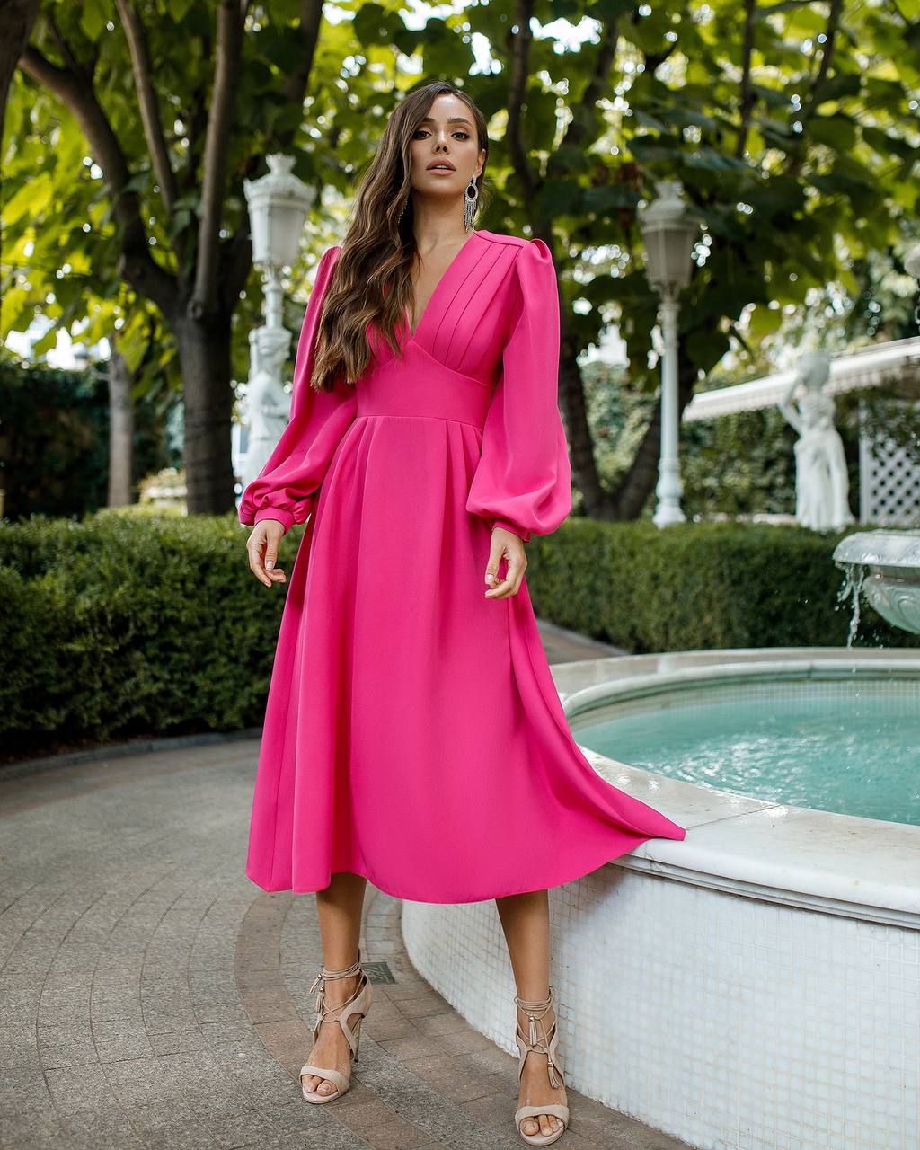 Woman in a bright pink dress standing by a pool with greenery in the background