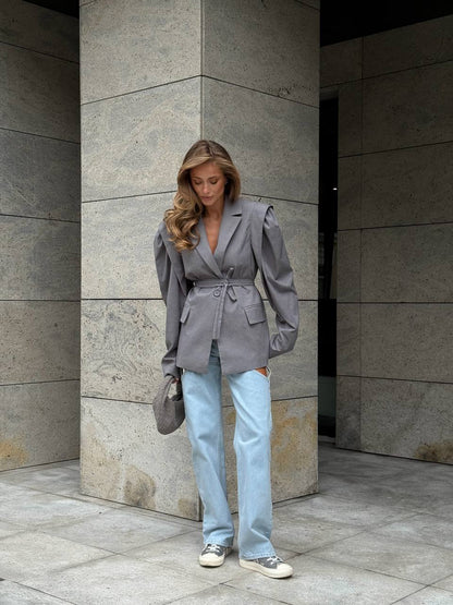 Woman wearing a gray blazer and light blue jeans standing against a stone wall.