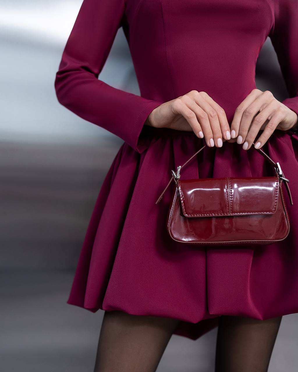 A close-up of a model wearing a deep magenta long-sleeve mini dress and holding a matching glossy patent leather handbag with silver-tone hardware.