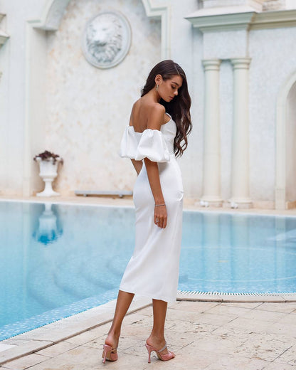 Woman in a white off-shoulder dress standing by a pool with classical architecture in the background