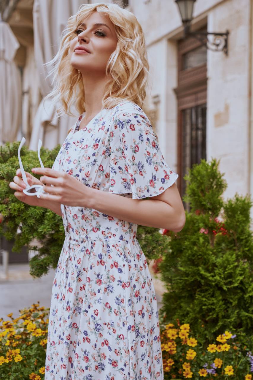 Woman in a floral dress standing outdoors with flowers and greenery around.