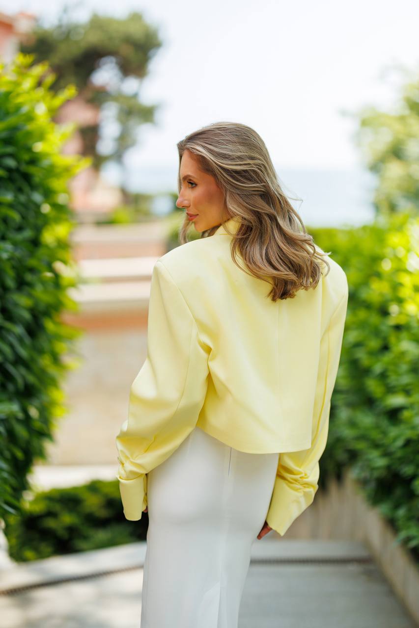 Woman wearing a yellow blouse and white pants in an outdoor setting with greenery.