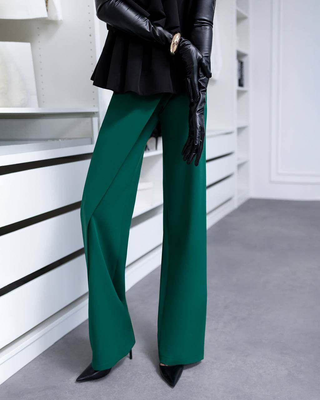 A model wearing emerald green wide-leg trousers, a black peplum top, black leather gloves, and black pointed-toe heels, standing in a modern white interior setting.