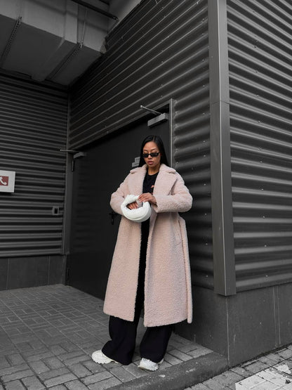 Person wearing a long pink coat standing against a dark wall.