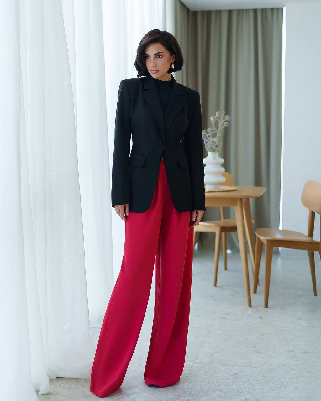 Woman wearing a black blazer and red pants in a room with white curtains and a wooden table.